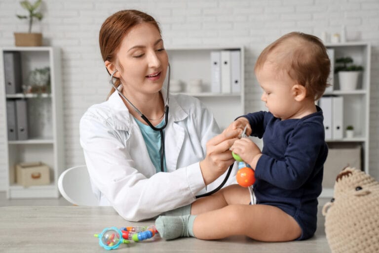 young happy female pediatrician with stethoscope examining cute little baby on table in clinic